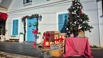 A festive outdoor Christmas scene featuring a decorated Christmas tree, wrapped gifts with red ribbons, a cheerful Santa mailbox, and a beautifully adorned blue door with holiday garlands.