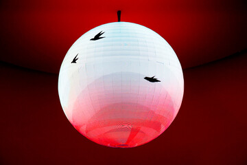 Digital globe. A large, glowing sphere suspended from above against a solid, dark red background. The sphere's surface has a digital or pixelated appearance.