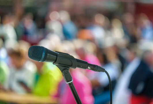 Seitliche Nahaufnahme eines Mikrofons im Halter vor einer Masse an unkenntlichen Menschen im Publikum im Hintergrund auf einer Open-Air Veranstaltung &ndash; selektiver Fokus, sehr viel Tiefenunsch&auml;rfe