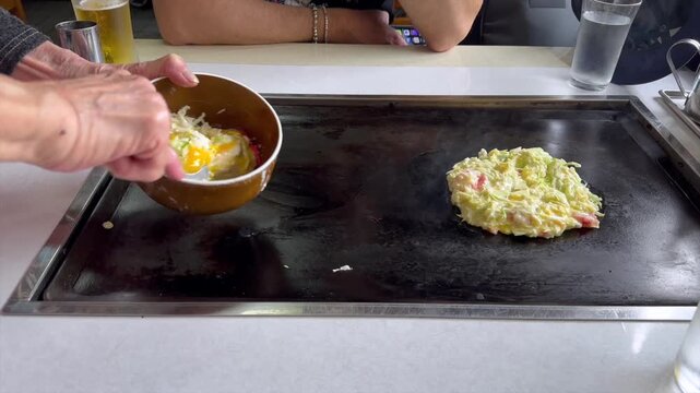 Okonomiyaki on teppan in a small restaurant in japan, an elderly chef pours batter and adds noodles and bacon for a second pancake filmed as part of cooking two portions