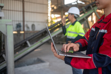 Worker Using Laptop in Industrial Setting with Colleague Talking on Walkie-Talkie in Background at Construction Site