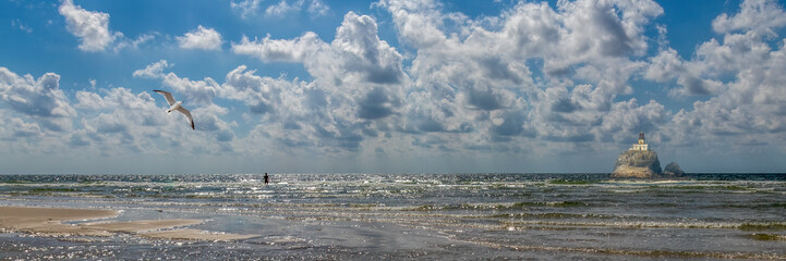 Seagull flying over ocean water near paddleboarder and old lighthouse under dramatic blue sky with copy space