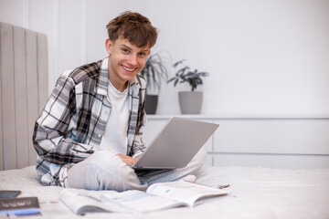 A teenage boy in a plaid shirt sits on his bed working on a laptop surrounded by textbooks and notebooks. A realistic scene of a high school student studying, doing homework, or attending online ©  Tsvyk L.
