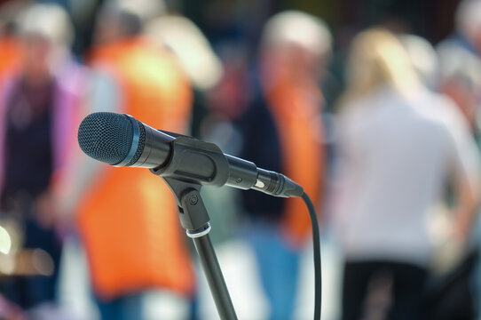 Seitliche Nahaufnahme eines Mikrofons im Halter vor einer Masse an unkenntlichen Menschen im Publikum im Hintergrund auf einer Open-Air Veranstaltung &ndash; selektiver Fokus, sehr viel Tiefenunsch&auml;rfe