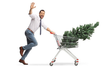 Cheerful man running with a christmas tree in a shopping cart and waving at camera