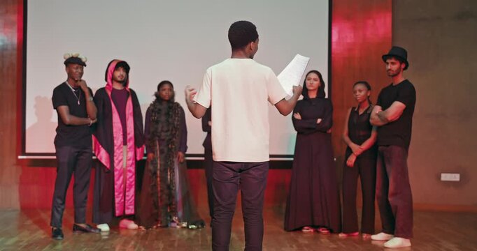 African young adult gen z male man hold scripts paper  read play story lines scene standing indoor stage hall. Happy group boy girl clap hand enjoy do work show drama practice day inside campus place
