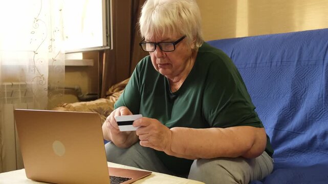 Senior woman sitting on sofa holding credit card and using laptop with product purchase at home. Concept of online banking, e-commerce, digital literacy, and internet payment