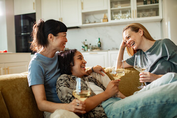 Adult friends laughing and drinking wine on home sofa