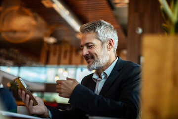Mature man smiling, using smartphone and drinking coffee at cafe