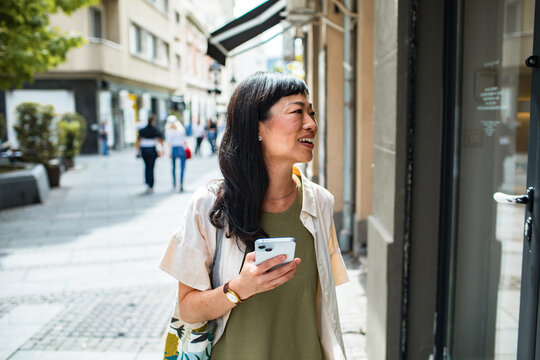 Adult woman smiling with smartphone while window shopping on city street