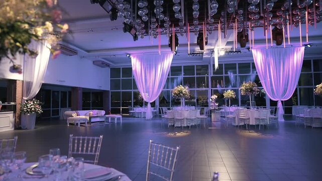 Modern glass‑walled ballroom set for wedding reception, with round tables dressed in white linens, tall floral centerpieces, chiavari chairs, soft purple uplighting on draped curtains and disco balls