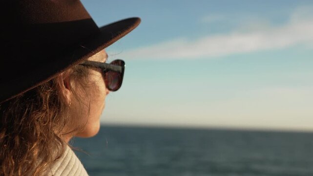 Woman, traveler, ocean. Stylish female wearing hat and sunglasses gazes at the sea horizon during peaceful sunset.