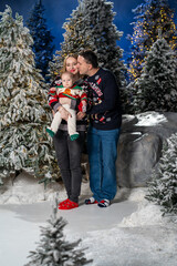 family Christmas moment with parents holding baby in snowy holiday setting. Both adults wear festive Christmas sweaters, while baby is dressed in a cute holiday outfit with Santa hat. Snowflakes fall 