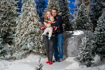 family Christmas moment with parents holding baby in snowy holiday setting. Both adults wear festive Christmas sweaters, while baby is dressed in a cute holiday outfit with Santa hat. Snowflakes fall 