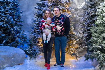 family Christmas moment with parents holding baby in snowy holiday setting. Both adults wear festive Christmas sweaters, while baby is dressed in a cute holiday outfit with Santa hat. Snowflakes fall 