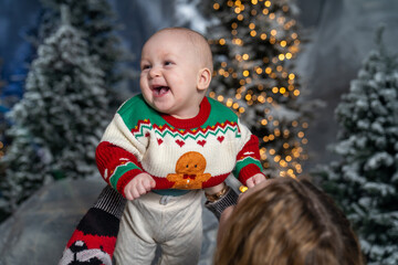mother lifts her smiling baby in a festive winter scene. The baby wears a colorful Christmas sweater with a gingerbread design, surrounded by snowy trees and warm holiday lights