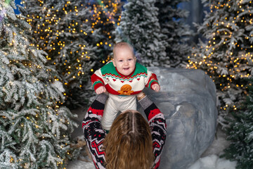 mother lifts her smiling baby in a festive winter scene. The baby wears a colorful Christmas sweater with a gingerbread design, surrounded by snowy trees and warm holiday lights