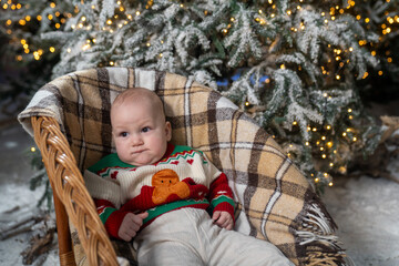 baby relaxing in a wicker chair with a cozy plaid blanket, wearing a festive Christmas sweater with a gingerbread design. Snow-covered Christmas tree branches and warm holiday lights create a magical 