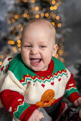 Close-up portrait of a laughing baby wearing a festive Christmas sweater with a gingerbread design. Warm holiday bokeh lights glow in the background