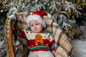 Baby in a Santa hat and festive Christmas sweater, gingerbread man,  sitting in a cozy wicker chair with a warm blanket, surrounded by snowy Christmas tree branches and twinkling holiday lights
