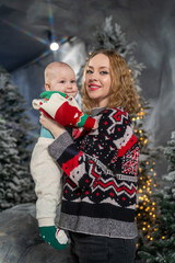 portrait of mother holding baby in winter wonderland setting. Both are dressed in cozy Christmas sweaters mother in  patterned style knit and baby in colorful holiday sweater with red, green, cream 