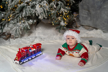 A cute baby in a colorful Christmas sweater and Santa Claus hat lies on the snowy ground, surrounded by warm festive lights on a train toy and a glowing Christmas tree