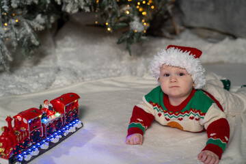 A cute baby in a colorful Christmas sweater and Santa Claus hat lies on the snowy ground, surrounded by warm festive lights on a train toy and a glowing Christmas tree