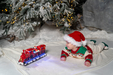 A cute baby in a colorful Christmas sweater and Santa Claus hat lies on the snowy ground, surrounded by warm festive lights on a train toy and a glowing Christmas tree
