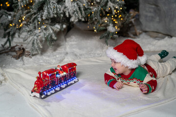 A cute baby in a colorful Christmas sweater and Santa Claus hat lies on the snowy ground, surrounded by warm festive lights on a train toy and a glowing Christmas tree