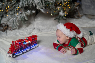 A cute baby in a colorful Christmas sweater and Santa Claus hat lies on the snowy ground, surrounded by warm festive lights on a train toy and a glowing Christmas tree