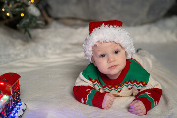 A cute baby in a colorful Christmas sweater and Santa Claus hat lies on the snowy ground, surrounded by warm festive lights on a train toy and a glowing Christmas tree