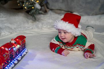 A cute baby in a colorful Christmas sweater and Santa Claus hat lies on the snowy ground, surrounded by warm festive lights on a train toy and a glowing Christmas tree
