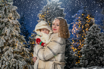 Mother holding baby outdoors in a snowy winter setting, surrounded by Christmas lights and trees. Mother is dressed in a warm beige winter coat, baby wears a cozy sweater and a fluffy cream hat