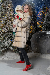 Full-body winter portrait of mother holding baby outdoors in snowy, festive setting. Mother is wearing a long beige puffer coat, black jeans, and red winter shoes, while baby is dressed in hooded hat