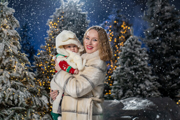 Mother holding baby outdoors in a snowy winter setting, surrounded by Christmas lights and trees. Mother is dressed in a warm beige winter coat, baby wears a cozy sweater and a fluffy cream hat