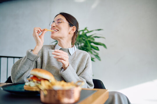 Young woman with glasses eating french fries and hamburger at a modern restaurant, having a happy and relaxing lunch