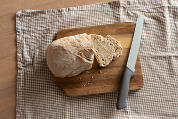 Sliced bread loaf with bread knife on cutting board on brown checkered kitchen towel in cozy home kitchen. Rustic kitchen atmosphere, home cooking, breakfast, bakery products, food preparation