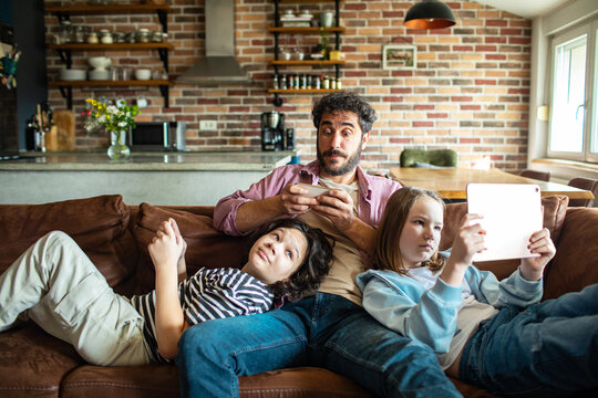 Adult father with children using devices on home sofa, relaxed