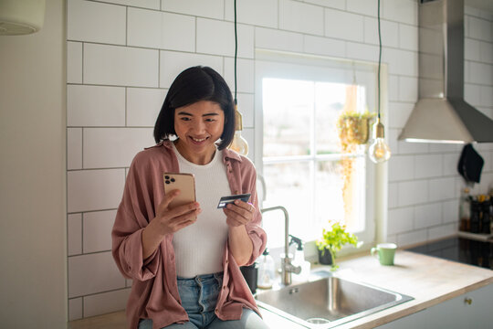 Young adult woman smiling using smartphone and card at home kitchen - Powered by Adobe