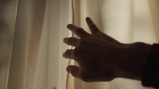 Hand draws open window curtain to reveal early morning sunlight shining in home. Close up on person's hand pulling drape or fabric inside house or apartment. Hope energy new solutions positive future