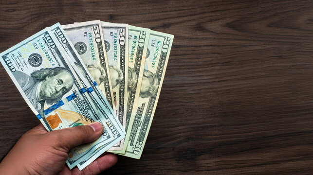 Unrecognizable person holding a stack of US dollar bills of various denominations on a dark wooden surface.