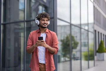 Young smiling Indian man wearing green headphones walking around the city, using his phone and looking to the side