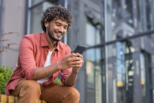 Smiling young Indian man sitting on a bench outside wearing headphones and using his mobile phone