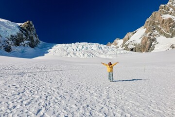 Mt Cook Glacier, NZ - May 24, 2025 (Glacier Landing): A person in a yellow jacket stands on vast snowfield, arms wide, framed by towering rock walls, a colossal glacier icefall. A moment of triumph. © Chonlatit