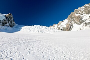 Obraz premium Mt Cook Glacier, NZ - May 24, 2025 (Glacier Landing): High-contrast, vast snowfield leading to a colossal, blue-tinged glacier icefall. Scene is dramatically framed by towering rock wall, blue sky.