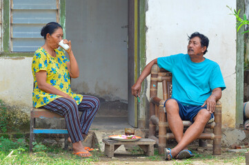 A middle-aged villager couple sits on wooden chairs on their terrace, relaxing after work, the father sits casually while the mother drinks from a white cup enjoying the break.