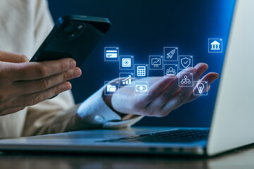 Hands using smartphone to interact with digital icons while working on laptop in a modern setting