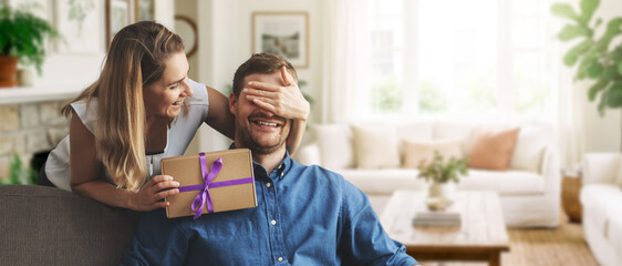woman giving surprise gift to beloved man at home. closed eyes with hand from behind. banner with copy space