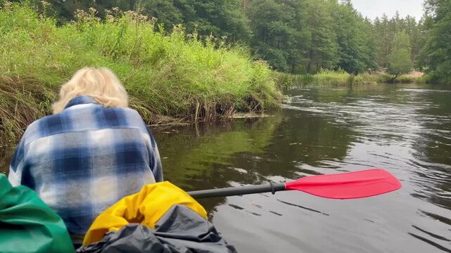 Woman resting in canoe as riverbank vegetation drifts past, holding snack while red paddle lies still beside her during calm Irbe River glide in Latvia