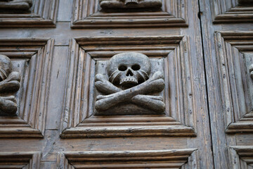 skull and crossbones carved into an old, weathered wooden door panel, evoking themes of memento mori, death, history, and gothic architecture
